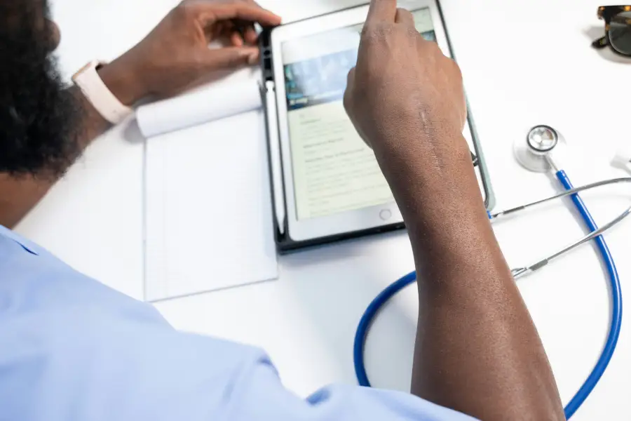 Medical professional in scrubs reviewing X-ray records on a tablet with a stethoscope and notepad on a white desk.