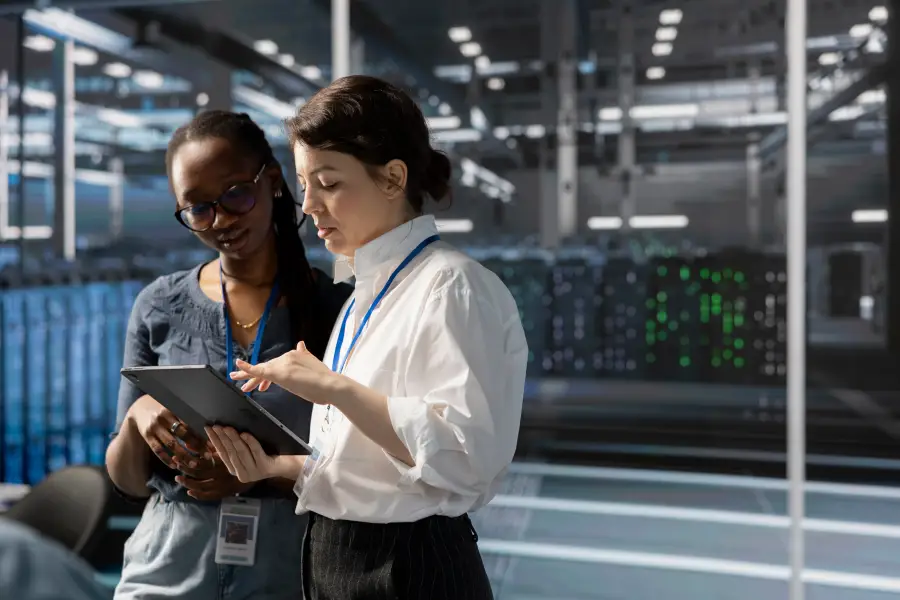 Two cleared female professionals reviewing data on a tablet inside a secure server facility with active equipment.