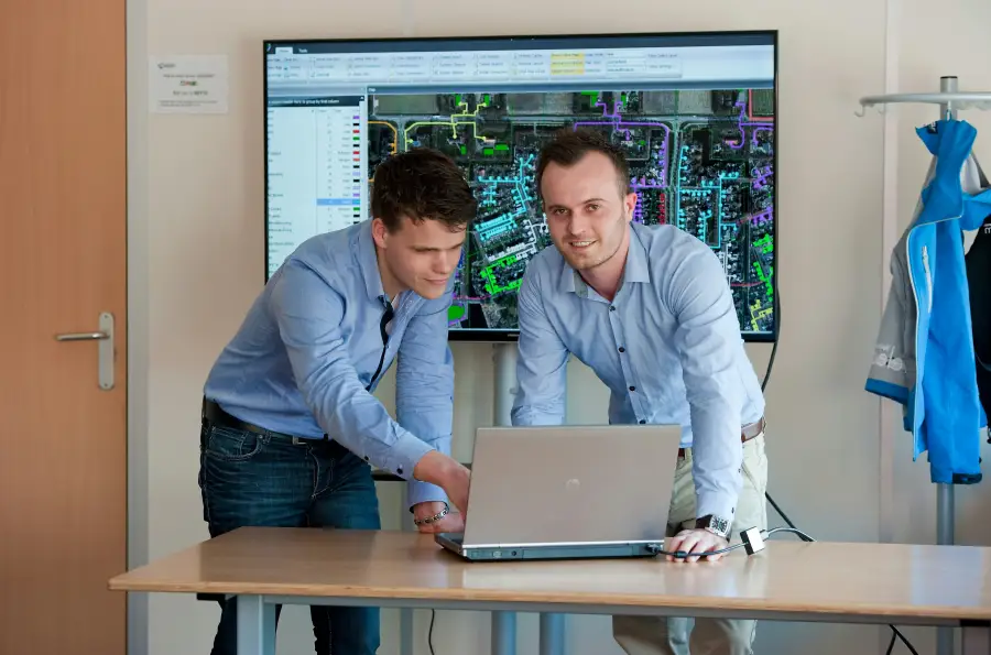 Two cleared professionals in button-down shirts review GIS security mapping software on a laptop with aerial network data