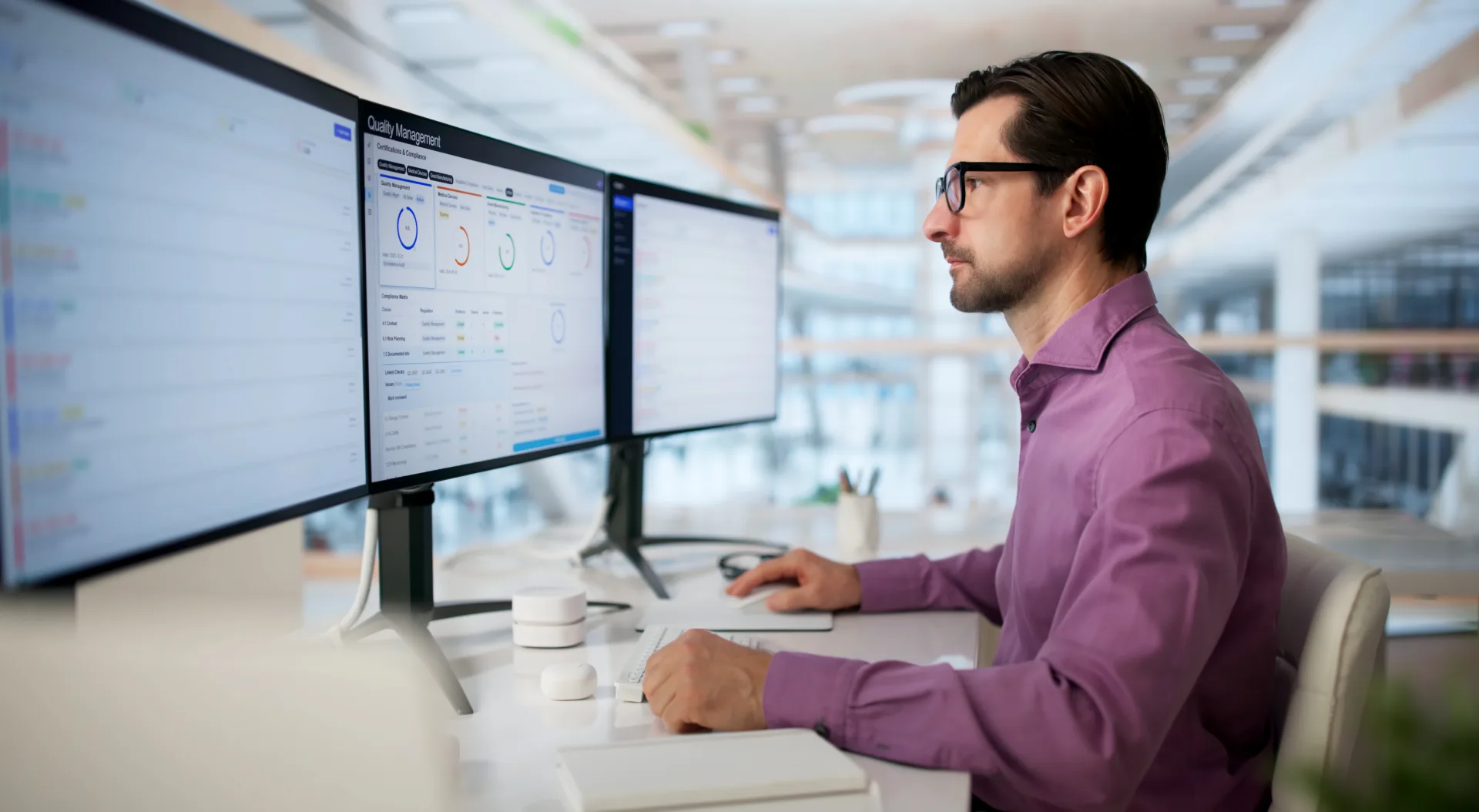 Cleared security professional reviews quality management and compliance dashboards across three monitors in a modern office.