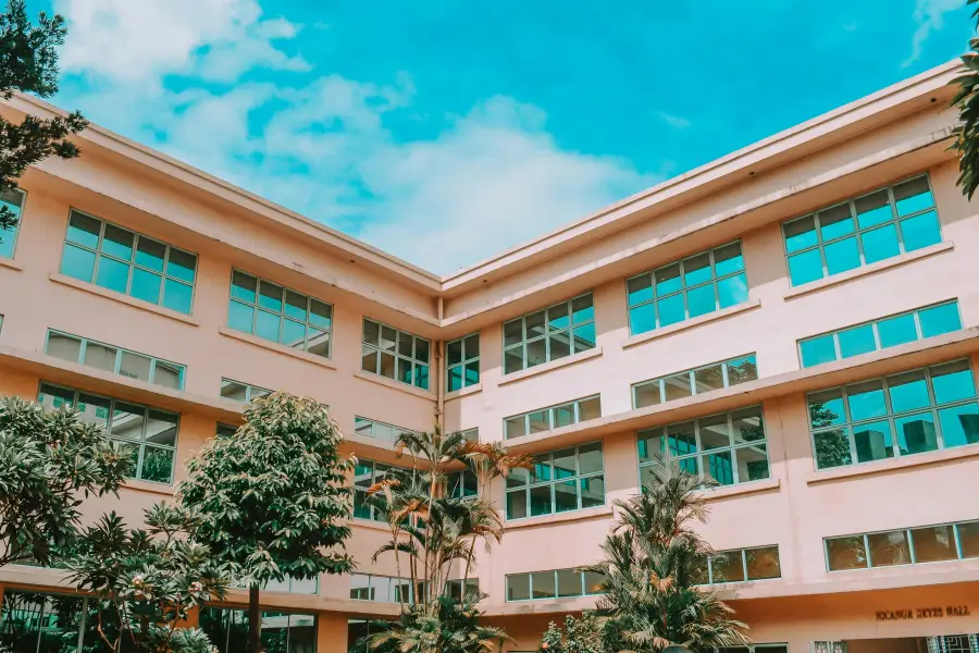 Exterior of Pacaños Reyes Hall; a multi-story peach-colored building with large windows surrounded by tropical trees.