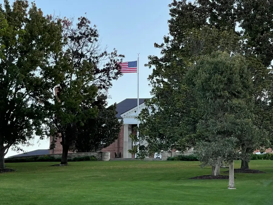 American flag flying at full staff in front of a brick colonial-style government building surrounded by mature trees and m