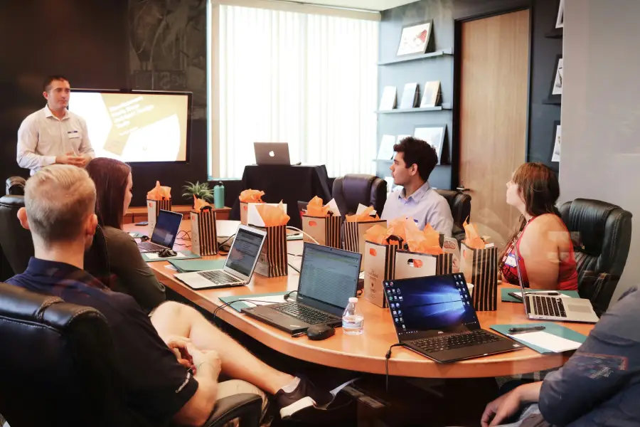 Presenter leads a security briefing for cleared personnel seated around a conference table with laptops and gift bags.