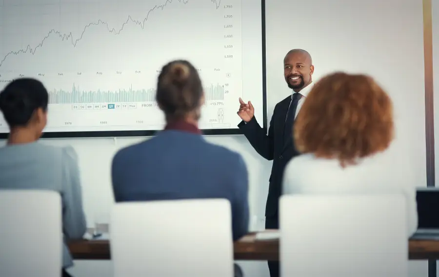 Suited presenter points to stock market chart on screen while three seated professionals attend a security briefing.