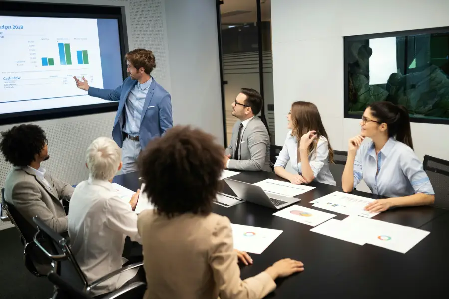 Business team reviews 2018 budget cash flow charts during a boardroom briefing led by a presenter at a display screen.