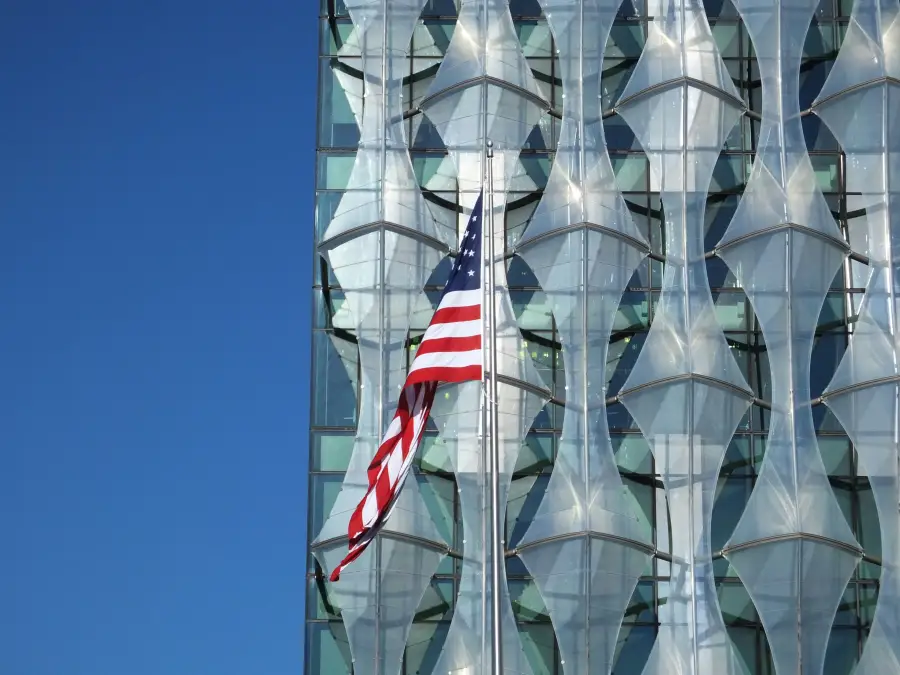 American flag flying in front of the modern glass-facade US Embassy building in London under a clear blue sky.