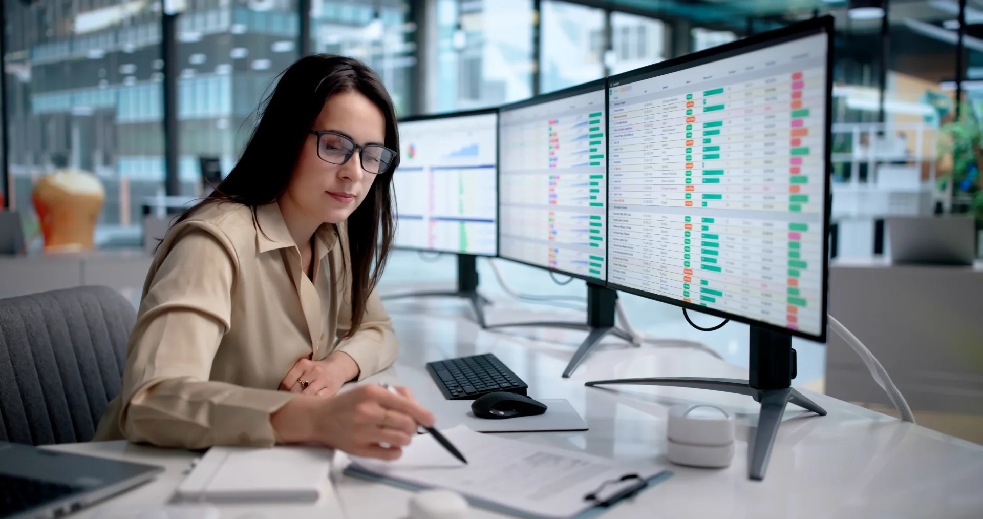 Woman in glasses reviews data dashboards on dual monitors at an enterprise office desk; taking notes with a pen.
