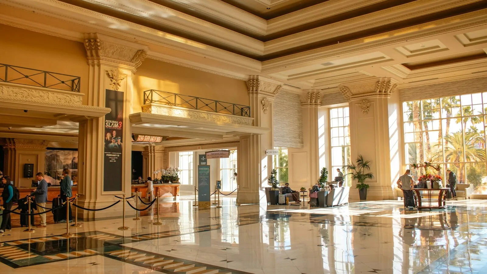 Guests crossing a sunlit hotel lobby past a staffed reception desk and a rope line.