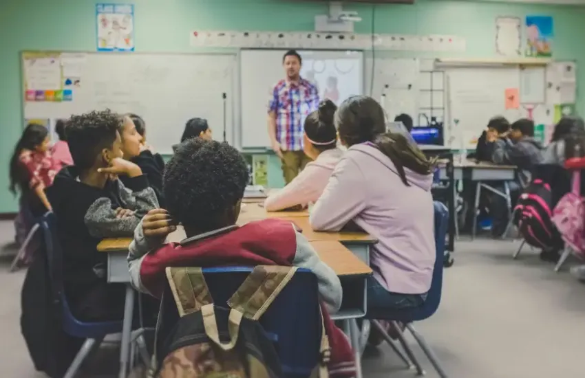 Students at desks face a male instructor presenting on a smartboard in a classroom security briefing setting.