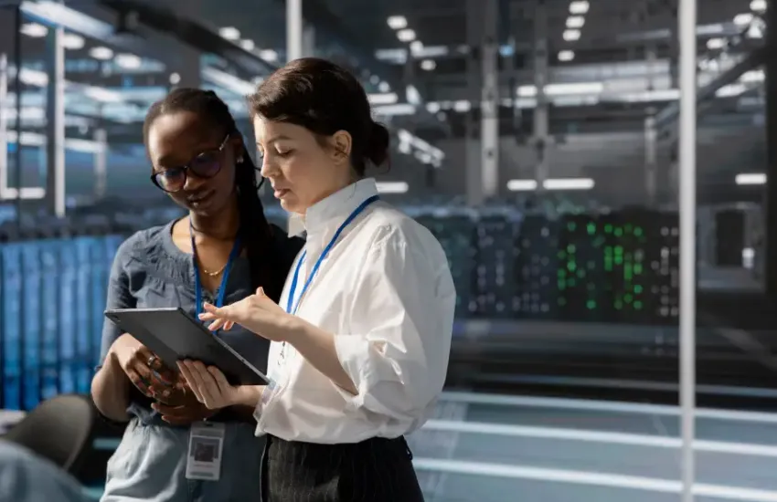 Two cleared female professionals reviewing data on a tablet inside a secure server facility with active equipment.