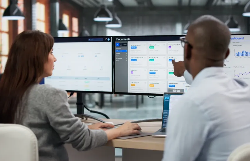 Two professionals reviewing classified documents and security dashboards on multiple monitors in a modern office environment.