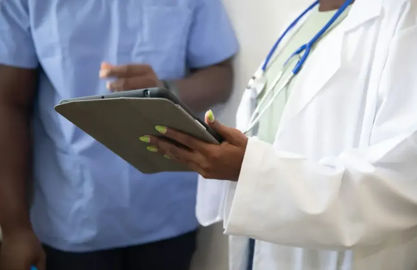 Doctor in white coat with stethoscope reviews tablet data alongside a colleague in blue scrubs in a medical setting.