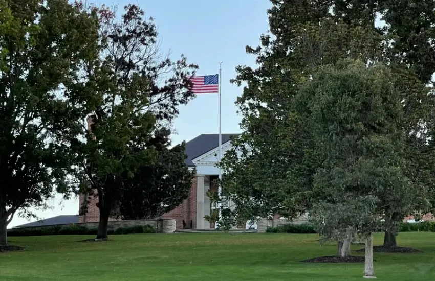 American flag flying at full staff in front of a brick colonial-style government building surrounded by mature trees and m...