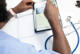 Medical professional in scrubs reviewing X-ray records on a tablet with a stethoscope and notepad on a white desk.