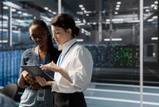 Two cleared female professionals reviewing data on a tablet inside a secure server facility with active equipment.