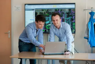 Two cleared professionals in button-down shirts review GIS security mapping software on a laptop with aerial network data...