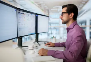 Cleared security professional reviews quality management and compliance dashboards across three monitors in a modern office.