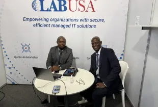 Two professionals in business attire seated at a LABUSA booth table with laptops; representing managed IT and AI security...