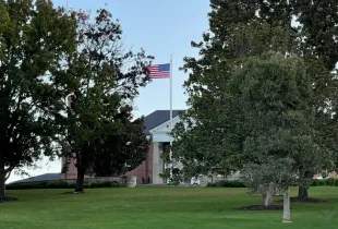 American flag flying at full staff in front of a brick colonial-style government building surrounded by mature trees and m...