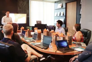 Presenter leads a security briefing for cleared personnel seated around a conference table with laptops and gift bags.