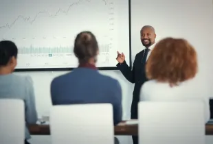 Suited presenter points to stock market chart on screen while three seated professionals attend a security briefing.