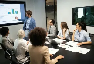 Business team reviews 2018 budget cash flow charts during a boardroom briefing led by a presenter at a display screen.