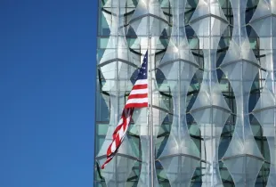 American flag flying in front of the modern glass-facade US Embassy building in London under a clear blue sky.