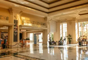 Guests crossing a sunlit hotel lobby past a staffed reception desk and a rope line.